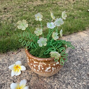 May include: A ceramic pot with a speckled brown and white design holds a beaded flower arrangement. The flowers are light green with green stems. Two white and yellow plumeria flowers rest on the stone surface.