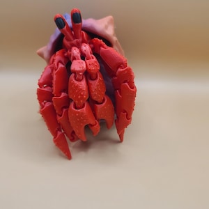May include: A red hermit crab ornament with detailed claws and eyes, emerging from a tan and purple shell. The figurine is set against a neutral background.