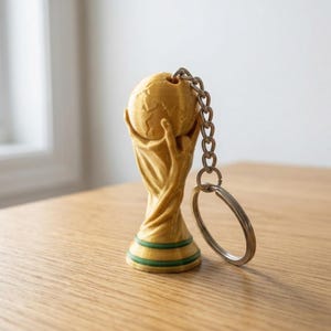 May include: A gold-colored keychain featuring a miniature trophy design with a globe held aloft by two figures. The base of the trophy has two thin green horizontal stripes. A silver-toned metal chain and a circular split ring are attached to the top.
