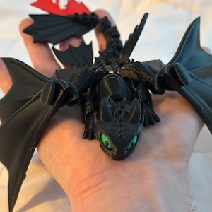 May include: A black dragon figurine with large wings and green eyes. The dragon has a detailed, textured surface and is posed with its wings spread. The figurine is held in a hand, showcasing its size and design.