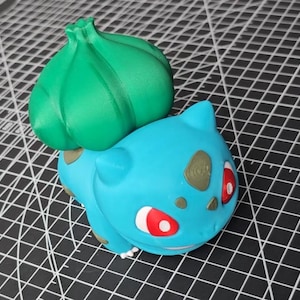 Puede incluir: Una figura de Bulbasaur impresa en 3D, un personaje de la franquicia Pokémon. La figura es principalmente azul claro con un gran bulbo verde en la espalda. Tiene ojos rojos y manchas marrones.