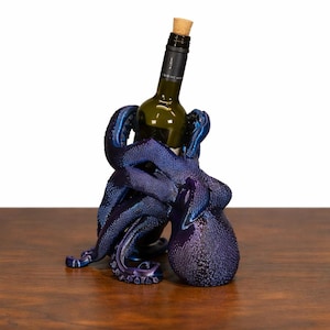 May include: A blue and purple octopus wine bottle holder. The octopus sculpture is holding a green wine bottle with a cork. The holder is on a wooden surface.