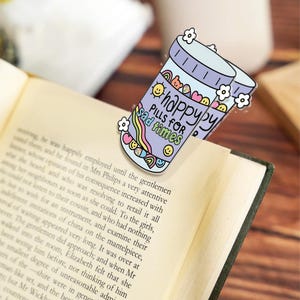 May include: A whimsical bookmark featuring a purple pill bottle design with the words "happy pills for sad times." The bookmark is adorned with flowers, rainbows, and smiley faces, adding a touch of cheerfulness to reading.