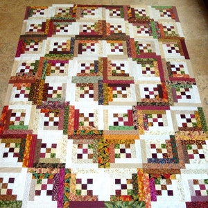 May include: A quilt with a log cabin pattern in shades of brown, green, and white. The quilt is made up of squares of different fabrics, each with a unique pattern.