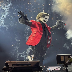 May include: A performer in a red sequined jacket with the text "FUCK YOU" on the sleeve, black pants, and a white face mask, singing into a microphone. The stage is lit with pyrotechnics and smoke.