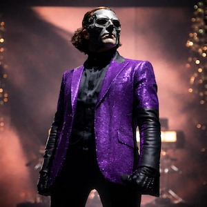 Op de afbeelding: Een persoon in een paarse pailletten blazer met zwarte leren mouwen, zwarte handschoenen en een zwarte broek. De persoon draagt ook een zilveren masker. De achtergrond is een podium met lichten.