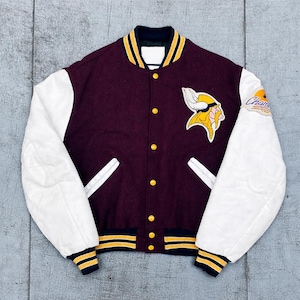 Puede incluir: Chaqueta universitaria granate y blanca con un parche con el logotipo de los Vikingos en amarillo y blanco. La chaqueta tiene mangas blancas, cierre de botones y ribetes a rayas amarillas y azul marino en el cuello, puños y dobladillo. La manga derecha tiene un parche de "Champions".