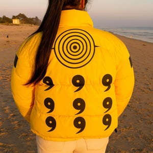 May include: A vibrant yellow puffer jacket featuring a black spiral design and the number "9" repeated. The jacket is shown on a beach. The back of the person wearing the jacket is visible.