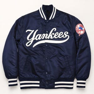 Puede incluir: Chaqueta de béisbol Yankees de satén azul marino con detalles blancos en el cuello, los puños y el dobladillo. La palabra "Yankees" está bordada en blanco en el pecho. Un parche con el logotipo de los Yankees está en la manga izquierda.