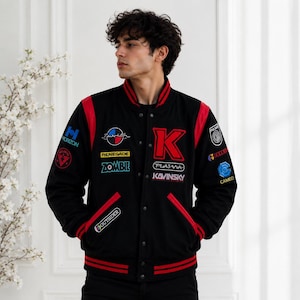 Könnte beinhalten: Eine schwarze Collegejacke mit roten Ärmeln und Besätzen sowie einer Druckknopfleiste. Die Jacke ist mit verschiedenen gestickten Aufnähern verziert, darunter ein großer roter Buchstabe K sowie die Schriftzüge Kavinsky, Plasma, Zombie und Renegade.
