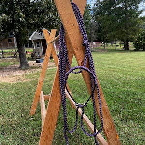 Puede incluir: Un cabestro y una cuerda de caballo trenzados de color púrpura se exhiben en un soporte de madera. El cabestro presenta una muserola de metal negro y una cuerda de plomo púrpura a juego. El soporte es de madera natural y está colocado al aire libre sobre una zona de césped.