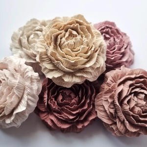 Può includere: Una collezione di fiori fatti a mano, simili alla carta, in varie tonalità di rosa, beige e crema. I fiori hanno petali dettagliati e stratificati, creando un aspetto realistico simile alla peonia. L'arrangiamento è ambientato su uno sfondo bianco.