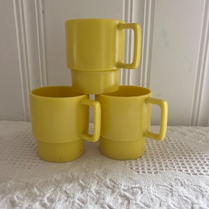 May include: Three yellow plastic mugs stacked together. Each mug has a handle and a stepped base. The mugs are a solid, bright yellow color and appear to be vintage or retro in style. The mugs are sitting on a white lace tablecloth.