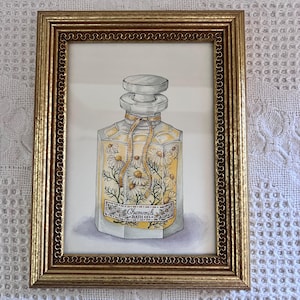 May include: A framed watercolor painting of a clear glass bottle filled with chamomile flowers. The bottle has a gold ribbon tied around the neck and a label that reads "Chamomile Bath Oil." The frame is gold with a decorative border.