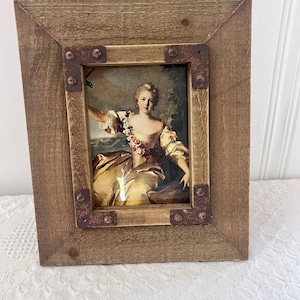 May include: A rectangular wooden picture frame with a portrait of a woman in a yellow dress. The frame has a rustic, weathered appearance with metal corner accents. The portrait features a woman with flowers in her hair, set against a landscape background.