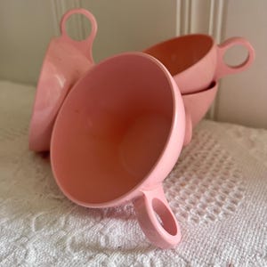 May include: Four pink plastic cups with rounded handles are stacked together. The cups are a light, solid pink colour and appear to be vintage. The cups are on a white, textured surface.