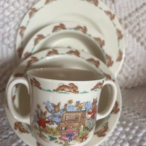 May include: A cream-colored children's dish set featuring a cup with two handles, a saucer, and two plates. The set is decorated with brown rabbits and a scene of rabbits playing. The plates and saucer have a border of running rabbits.