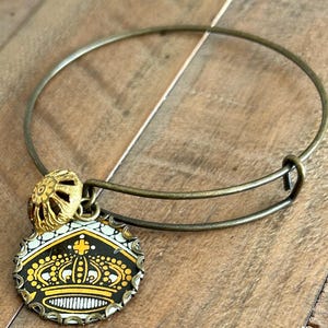 May include: An antique brass-colored bangle bracelet with a circular charm featuring a gold crown design on a black and white background. The charm is attached to the bracelet with a small, decorative gold bead.