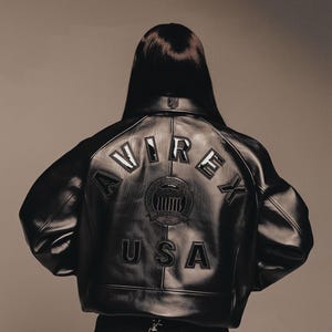 Könnte beinhalten: Schwarze Lederjacke mit dem Wort "AVIREX" auf dem Rücken, über einem kreisförmigen Emblem und dem Wort "USA". Die Jacke hat einen klassischen Bomberstil mit geripptem Kragen und Bündchen.