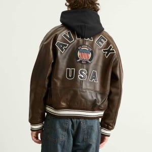 May include: Brown leather jacket with a black hood. The back of the jacket features white lettering that spells out "AVIREX" and "USA". A patch with the American flag is also visible.