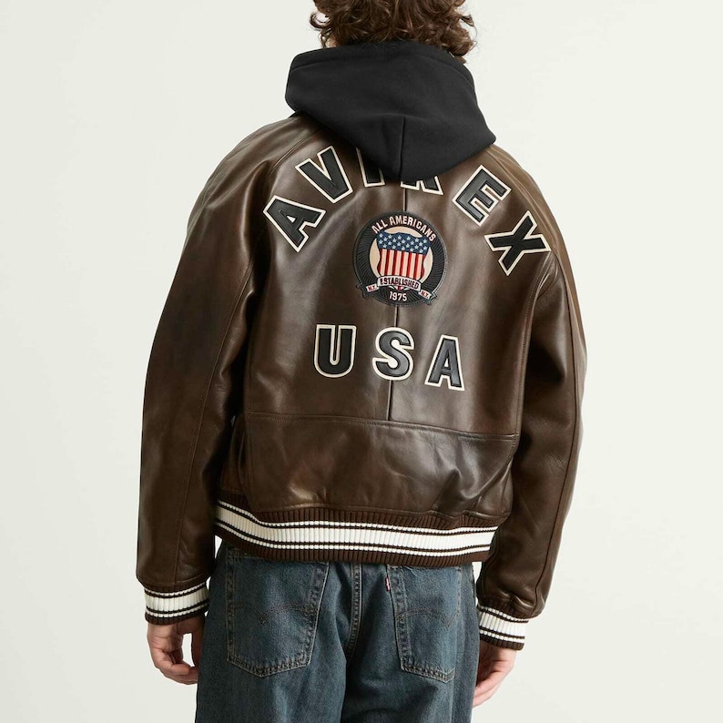 May include: Brown leather jacket with a black hood. The back of the jacket features white lettering that spells out "AVIREX" and "USA". A patch with the American flag is also visible.