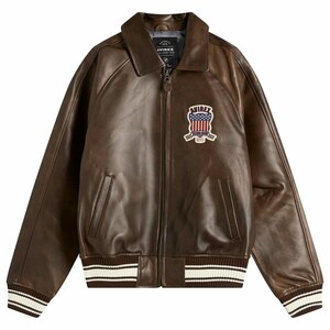 May include: Brown leather bomber jacket with a zip-up front, a collar, and a patch on the chest. The jacket has white and brown striped cuffs and waistband. The patch features a red, white, and blue design with the text "AVIREX".