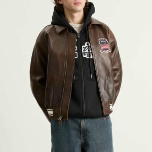 May include: A brown leather jacket with a zip closure, a collar, and white stripes on the cuffs and hem. The jacket features a patch with text and an American flag design on the upper left chest. The person is wearing a black hoodie underneath.