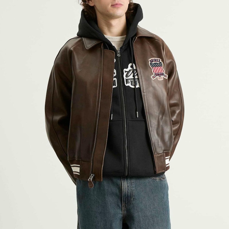 May include: A brown leather jacket with a zip closure, a collar, and white stripes on the cuffs and hem. The jacket features a patch with text and an American flag design on the upper left chest. The person is wearing a black hoodie underneath.