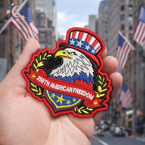 May include: Embroidered patch featuring an American bald eagle wearing a patriotic top hat. The patch includes the text "250th AMERICAN FREEDOM" on a red banner, with a blue and gold shield design. The patch is red, white, blue, black, and yellow.