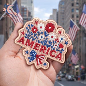 May include: Embroidered patch with the word "AMERICA" in red, surrounded by red, white, and blue floral designs and stars. The patch has a beige border and a blue and red bow. The background features American flags.