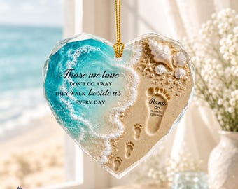 May include: A heart-shaped glass ornament featuring a beach scene with turquoise ocean waves, sandy shore, seashells, and a starfish. It displays the text "Those we love DON'T GO AWAY THEY WALK beside us EVERY DAY." and a footprint with "Nana".