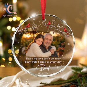 May include: A round, faceted glass memorial ornament with a red ribbon hanger. It features a photo of a person hugging an older man, red cardinals on snowy branches, and the text: Those we love don't go away They walk beside us every day Dad 1935-2026.