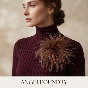 May include: A dark burgundy ribbed turtleneck sweater featuring a large, circular brown feather brooch pinned to the chest. The brooch is composed of numerous thin, radiating feathers that create a voluminous, textured, and spherical appearance.