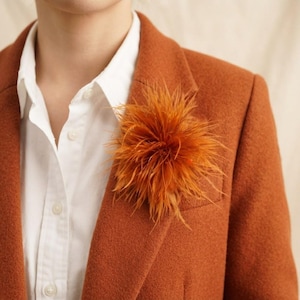 May include: A vibrant orange feather brooch shaped like a round, textured pom-pom. The accessory is pinned to a rust-colored blazer worn over a crisp white button-down shirt. The feathers are fine, wispy, and radiate outward from the center.