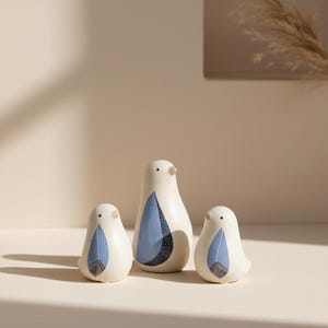 Può includere: Tre statuette di uccelli in ceramica di diverse dimensioni. Gli uccelli sono bianchi con disegni di piume blu e nere. L'uccello più grande ha un'ala blu a fantasia. Le statuette sono esposte su una superficie chiara.
