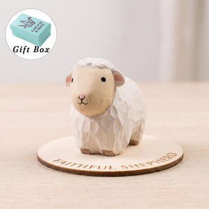 May include: A hand-carved wooden sheep figurine with a cream-coloured face and white textured wool body, standing on a circular wooden base engraved with the text FAITHFUL SHEPHERD. A light blue gift box with an origami crane icon is shown above.