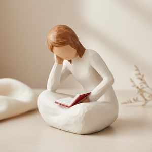 Op de afbeelding: Een beeldje van een zittende persoon in een witte jurk, die een boek met een rode kaft leest. De figuur heeft bruin haar en rust met haar hoofd op haar hand. Het beeldje is gemaakt van een lichtgekleurd materiaal.