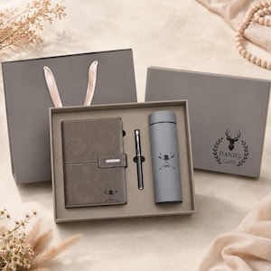 May include: A gray gift set containing a notebook with a strap closure, a pen, and a cylindrical bottle. The notebook and bottle feature a graphic of crossed arrows, a top hat, and a mustache with the letters K and T. The box lid displays a deer head.