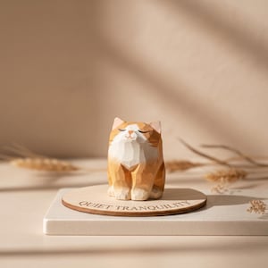 May include: A wooden cat figurine with orange and white coloring, sitting on a wooden platform. The cat has closed eyes and a smiling expression. The platform has the words "QUIET TRANQUILITY" engraved on it.
