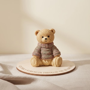 Op de afbeelding: Een beige teddybeerbeeldje met een veelkleurige trui, zittend op een houten voet. De voet is gegraveerd met de tekst "CUDDLY STRENGTH THROUGH THE SEASONS". De beer heeft een lieve uitdrukking.