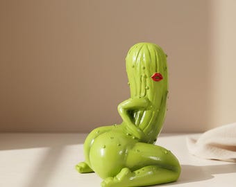 May include: A whimsical, green cactus figurine with red lips. The sculpture is posed in a reclining position, with one arm resting on its chest. The figurine has a smooth, glossy finish and is dotted with small bumps.