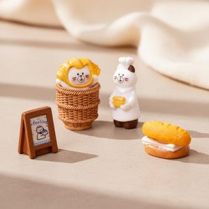 Könnte beinhalten: Eine Miniatur-Bäckerei-Szene mit einer Katzenfigur in einem Weidenkorb, einem Kochkater, der Brot hält, einem Sandwich und einem kleinen Holzschild mit dem Wort "feeling". Die Figuren sind in Braun-, Weiß- und Gelbtönen gehalten.