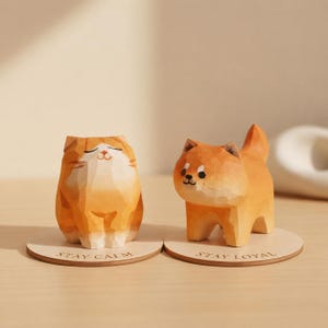 May include: Two wooden animal figurines: an orange cat and a dog. The cat has closed eyes and the text "STAY CALM" is on its base. The dog has a small tail and the text "STAY LOYAL" is on its base. Both are on a light brown surface.