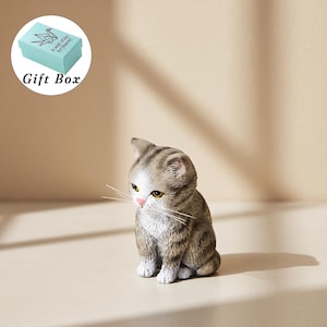 Op de afbeelding: Een klein, realistisch beeldje van een zittend cyperse kitten met grijs en bruin gestreepte vacht, een witte borst en een roze neus. Het kitten heeft gele ogen en lange, dunne witte snorharen. Een lichtblauw cadeau doosje staat linksboven.