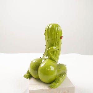 Peut inclure: Une sculpture de cactus verte fantaisiste avec une finition brillante. Le cactus est représenté dans une pose assise, avec des formes arrondies et de petits détails. Il a une paire de lèvres rouges et repose sur une base carrée de couleur claire.