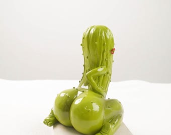 May include: A whimsical, green cactus sculpture with a glossy finish. The cactus is depicted in a seated pose, with rounded forms and small details. It has a pair of red lips and is resting on a square, light-colored base.