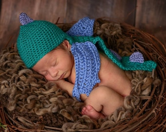 May include: A newborn baby sleeping in a woven nest, wearing a green and blue crocheted dinosaur hat and matching wings.