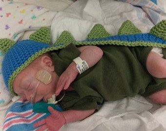 May include: A newborn baby wearing a blue and green crocheted dinosaur hat and a green onesie. The baby is lying on a white sheet with a pink and white striped blanket underneath.