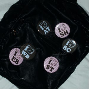 Op de afbeelding: Een verzameling ronde speldjes met engelenvleugel- en halo-ontwerpen. De speldjes tonen de tekst "I AT", "I SH", "I SC", "I ES" en "I ST" in verschillende kleuren, tegen een gestreepte en effen achtergrond. De speldjes zijn geplaatst op een zwart leren oppervlak.