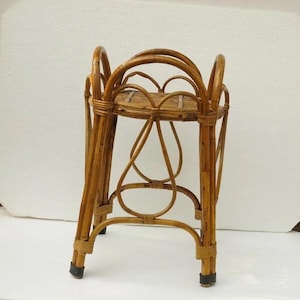 Könnte beinhalten: Ein Vintage-Pflanzenständer aus natürlichem Bambus. Der Ständer hat eine runde Oberseite, dekorative geschwungene Stützen und ein gewebtes Regal. Der Bambus hat eine warme, hellbraune Farbe und der Ständer hat einen klassischen, böhmischen Stil.